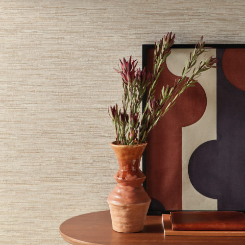 An image of a wooden side table featuring a brown ceramic vase filled with dried flowers, along with two brown notebooks and a painting of geometric shapes in rust, white and navy. The table is positioned against a wall on which Omexco’s Loom Stories wallcoverings are installed and is illuminated by natural light from a nearby window. This elegant linen and cotton weave is shown here in a soothing shades of beige and light brown. The combination of organic and geometric elements creates an interesting visual contrast, while the warm brown color scheme creates a cozy and welcoming ambiance.