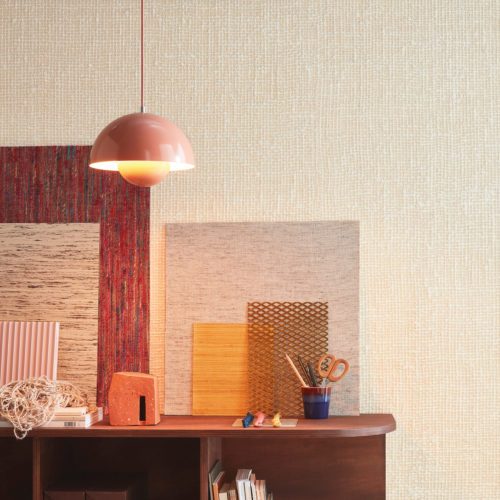 Interior designer's or architect’s inviting home office with a massive wooden shelving unit and pink vintage Flowerpot VP1 Hanging Lamp. We see samples of Omexco wallcoverings and other materials, books and small storage boxes laying on and in the shelving unit. On the wall, Omexco’s textile wallcovering collection “Loom Stories” is installed: a very textured weave of thick, off white yarns. A soothing neutral wall you would love to touch.