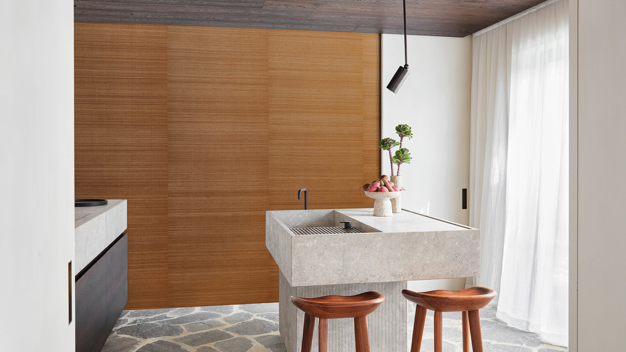 A bright and airy kitchen featuring a natural stone floor that adds a touch of earthy elegance. The walls are adorned with a stunning burnt orange silk wallcovering from Omexco’s textile collection ‘Loom Stories’. In the center of the room, there is a beautiful kitchen island made of stone, with a bowl of fresh vegetables and a vase with flowers on top. Two wooden barstools are placed around the island, providing comfortable seating for family and friends. The combination of natural materials, vibrant colors and functional design makes this kitchen a warm and inviting space for cooking and gathering.