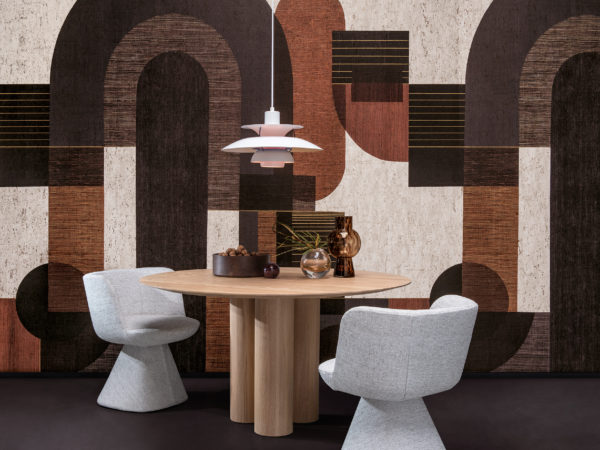 Example of a dining room with a bold graphic wallcovering showing a pattern of geometric shapes in grey, black, burnt orange and white hues. This panoramic design is digitally printed on cork and belongs to Omexco’s wallcovering collection “Casca”. We see a round wooden dining table in the center of the room with a white pendant lamp hanging above it. Decorative elements such as small glass vases and a bowl filled with walnuts are placed on the tabletop. Two soft grey dining chairs are positioned around the table. The overall aesthetic is modern with a touch of warmth and texture.