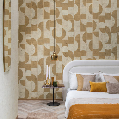 An image of a stylish bedroom featuring a light herringbone floor, a white bed with pillows and an ochre blanket, a mirror and a modern gold pendant lamp. A black marble side table provides a sleek contrast to the light colors, while a white and yellow geometric wallcovering adds a playful pop of pattern. The mosaic of contrasting geometric shapes is made of cork and belongs to Omexco’s wallcovering collection “Casca”. The overall effect is a fresh and modern space that is both inviting and visually intriguing.