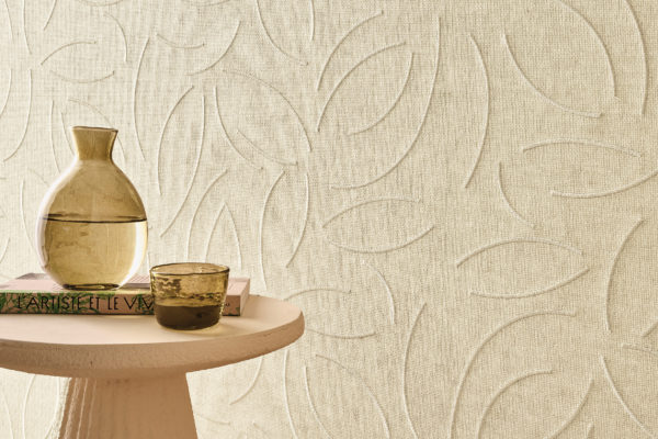 This serene interior highlights Omexco’s OAS221 reference, Verano, from the Oase collection. The wallcovering, crafted from exquisite paperweave, features delicate embroidered patterns inspired by organic forms, adding a sense of movement and sophistication to the space. The soft, neutral tones create an inviting and calming atmosphere, perfectly complemented by a sculptural side table, a glass water carafe, and a textured ceramic cup. The interplay of light and shadow on the embossed surface enhances its depth, making it a refined choice for elegant and natural interiors.