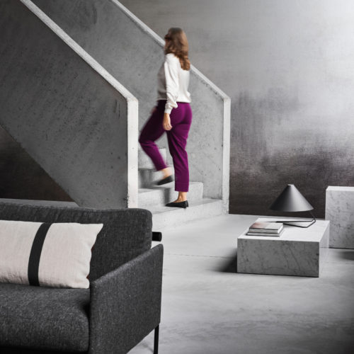 This striking interior features Omexco’s LAN292 reference, Evening Glow, from the Light at Night collection. The wallcovering showcases a captivating chiaroscuro effect, with a seamless gradient that transitions from deep shadow to soft illumination, evoking the beauty of twilight. The moody yet sophisticated ambiance is enhanced by raw concrete architectural elements, a minimalist marble coffee table, and a contemporary black lamp. A plush charcoal-grey sofa with a contrasting cushion adds warmth and comfort to the space, while a woman in vibrant magenta trousers ascends the concrete staircase, adding a dynamic touch to the scene.