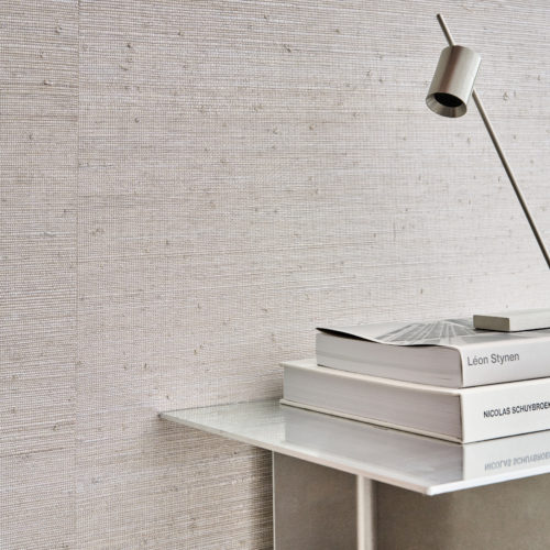 This minimalist interior features Omexco’s LAN201 reference, Cosmic, from the Light at Night collection. The wallcovering, crafted from natural-hued raffia, is delicately interwoven with metallic threads, creating a refined interplay of texture and shimmer. The neutral color palette complements the sleek, modern aesthetic of the space, where a brushed metal side table holds a carefully curated selection of architectural books. A sculptural reading lamp adds a touch of contemporary elegance, enhancing the serene and sophisticated atmosphere of this setting.