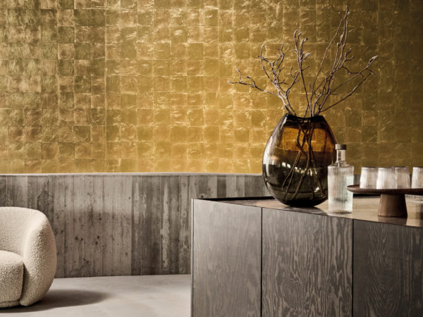 This striking interior highlights the LAN241 reference, titled Solar, from Omexco’s Light at Night collection. The wallcovering, crafted with gold leaf, showcases an intricate geometric pattern that plays with light, creating a rich and dynamic surface. The golden hues shimmer subtly, adding depth and warmth to the space. The combination of raw concrete walls and dark wood furniture provides a modern contrast, allowing the luxurious gold-leaf finish to take center stage. A sculptural vase with delicate branches enhances the natural elegance of the setting, while a cozy boucle armchair invites relaxation in this refined environment.
