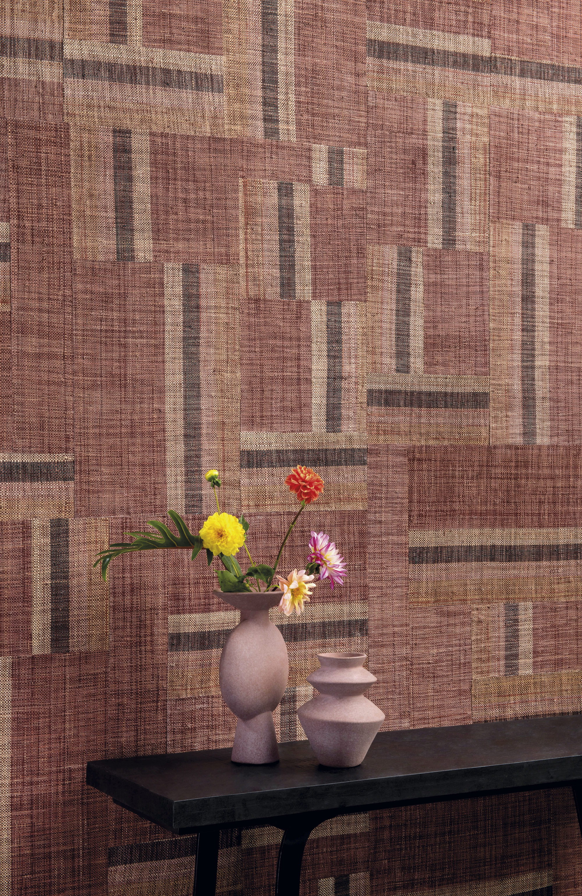 Authentic craftmanship embodies the bold Atelier collection designed by the creative minds of Omexco. This image shows an impressive red-brown patchwork of handwoven raffia. The perfect backdrop for a wooden sidetable with two pink ceramic vases and some field fresh flowers.