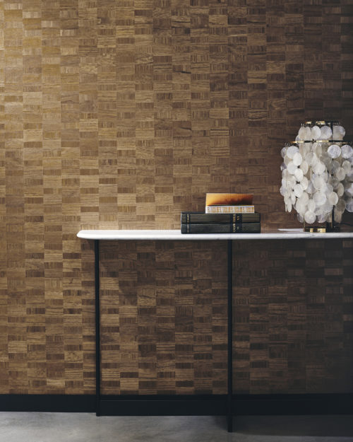 A decor shot featuring a marble side table against a wall adorned with Omexco's Imperialis marquetry wallcoverings. On the table, a collection of books is neatly arranged alongside an elegant Verpan capiz shell table lamp, casting a warm glow. The wallcoverings, crafted from paulownia wood, exhibit a striking design of contrasting rectangles, highlighting the natural beauty of the material