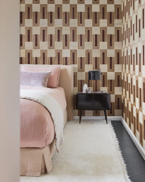 A bedroom with a pink bed atop a soft white carpet, complemented by a sleek black nightstand and table lamp. The floor contrasts with dark grey parquet. On the wall, Omexco's marquetry wallcovering collection "Imperialis" is displayed, showcasing a delicate interlocking pattern of wood against a metallic background. The shapes are brown, burgundy, and beige, creating a sophisticated and harmonious atmosphere.