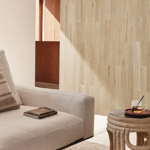 A serene living room corner featuring a beige sofa adorned with a striped pillow and topped with a stack of books. A beige carpet adorns the floor below. Next to the sofa sits an organic-shaped beige round side table, a design by Pascale Degueldre and Jehanne Hupin. The backdrop showcases Omexco's exquisite wood veneer wallcovering collection Imperialis, featuring a parquet-like natural wood design.