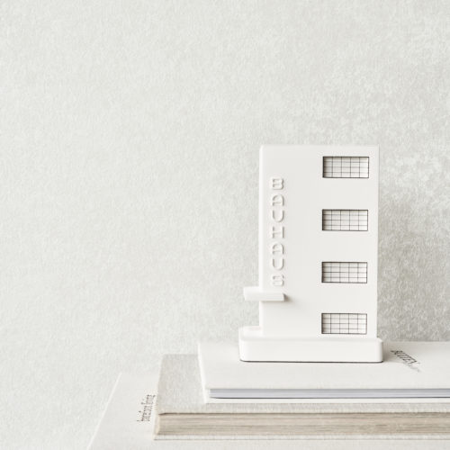 Decor Closeup of a pile of architecture books with a decorative sculpture designed by Chisel & Mouse that reproduces the famous Bauhaus Dessau building in small model. As a backdrop, we see Omexco's Edition collection, a carefully curated range of the brand’s most iconic faux-plain designs, reinterpreted in fresh colourways and complemented with appealing new textures. The white wallcovering is elaborated with a matte finish. This sophisticated patina-effect print discretely reflects the incoming light.