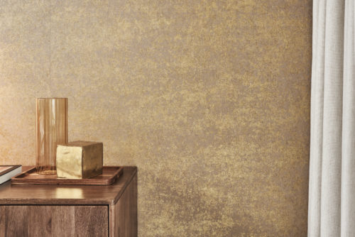 A corner of a room featuring a patina-effect wallcovering from Omexco's printed non-woven collection "Edition." The wall has a textured gold and brown finish. In the foreground, a wooden cabinet holds a decorative tray with a tall amber glass vase and a gold-colored cube. On the right, there is a glimpse of light-colored curtains.