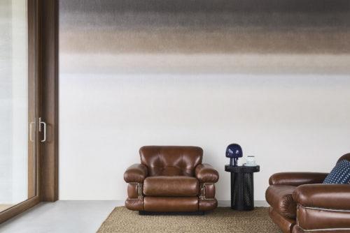 A stylish living room featuring a gradient printed non-woven wallcovering from Omexco's "Edition" collection. The wall transitions from dark gray and brown at the top to light beige at the bottom. The room includes a brown leather armchair, a matching sofa with a blue patterned cushion, and a black side table with a blue lamp and a tea set. A large window with wooden framing is on the left, and a beige rug covers the floor.