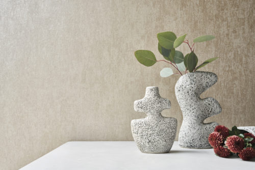 A minimalist setting featuring a plaster-like printed non-woven wallcovering from Omexco's "Edition" collection. The wall has a textured beige appearance. In the foreground, there are two uniquely shaped speckled vases, one containing green leaves. To the right, there are red flowers placed on a light-colored surface, enhancing the modern and artistic look of the space.