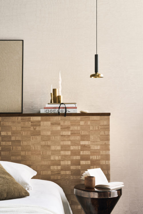 A sophisticated bedroom setting featuring Omexco's Pure collection, reference PU391, composed of textile warps with an elegant moiré effect. The wallcovering’s subtle relief enhances its texture, adding depth and a touch of understated luxury. The scene showcases a wooden bedside table with a rich dark finish, adorned with an open book and a wooden mug. To the left, a checkerboard wood headboard complements the natural tones, topped with stacked books, gold decorative candlesticks, and a sculptural twisted candle. Overhead, a pendant light with a brass accent provides a warm, inviting glow, perfectly harmonizing with the refined ambiance of the space.