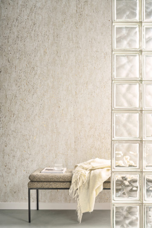 An elegant and serene space featuring Omexco's Pure collection, reference PU271, a wallcovering named Nebbiosa that highlights the subtle plain texture and the unique grain of cork with shimmering delicacy. The natural tones of the wallpaper add a soft, organic ambiance to the room, enhanced by the reflective glass block partition that introduces additional light and texture. The scene includes a modern bench with a textured fabric seat, adorned with a cream-colored throw and a neatly placed book with a glass tumbler on top. This harmonious composition celebrates natural materials and understated elegance, ideal for contemporary and minimalist interiors.