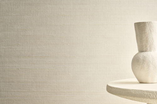 A close-up of a textured wallcovering from Omexco's Pure collection, reference PU462, featuring a weave of irregular sisal yarn in natural shades, set against a softly textured base. This sophisticated wallcovering brings an organic warmth to interiors, perfectly balancing rustic charm with modern elegance. On the right, a sculptural white vase rests on a round textured table, complementing the nature-inspired aesthetic.