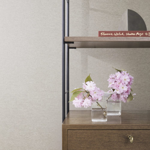 A close-up of a shelf in a minimalist room with a printed non-woven wallcovering from Omexco's "Edition" collection, featuring an intricate stripe pattern that blends smoothly into a relief texture, subtly evoking the weft of fabric. The wall has a textured beige appearance. On the wooden shelf, there are two glass vases with pink cherry blossoms. Above the shelf, there is a book titled "Florence Welch Useless Magic" and a geometric decorative object.