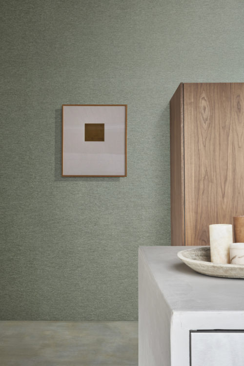 Home decor shot witha concrete kitchen counter in the front with a stone plate and marble vases on top. On the right, a  dark wooden  cabinet and on the left minimalist framed wall art. On the wall, we see a green wallcovering from Omexco’s Edition collection, a carefully curated range of the brand’s most iconic faux-plain designs. The intricate stripe pattern of the wallcovering blends smoothly into a relief texture, subtly evoking the weft of fabric.