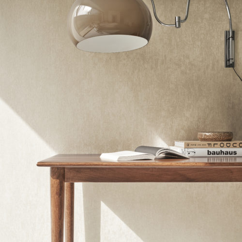 Home decor shot of a mid-century modern wooden table placed against a wall and some architecture books on top. 1960s vintage globe wall lamp withh a chrome adjustable wall fixture. On the wall, a beige/soft pink wallcovering from Omexco’s Edition collection, a carefully curated range of the brand’s most iconic faux-plain designs. The rich, plaster-like texture of the wallcovering conveys structure and serenity to every space.
