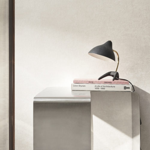 Closeup of a minimalist metal side table with a stack of architecture books and a sleek black table lamp on top. In the background is Omexco's Edition collection, featuring iconic faux-plain designs in fresh colors and new textures. The off-white wallcovering has subtle geometric patterns enhancing the semi-plain design.