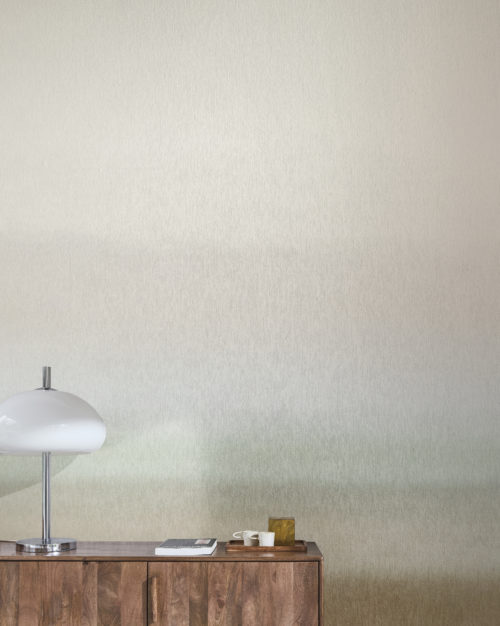A minimalist interior featuring a gradient printed non-woven wallcovering from Omexco's "Edition" collection. The wall transitions smoothly from light beige at the top to a darker beige at the bottom. In the foreground, there is a wooden cabinet with a white lamp, a book, and a tray with cups and a small decorative item. The overall aesthetic is clean and modern.