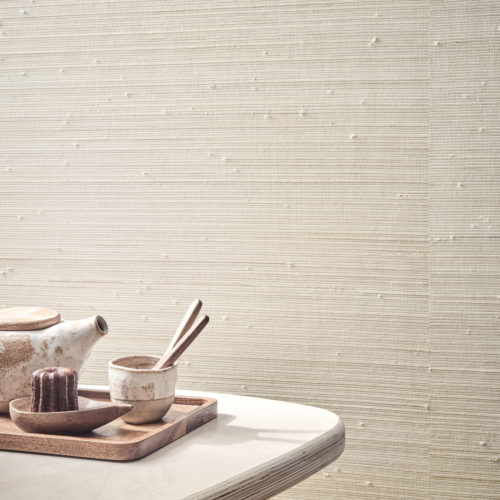 A serene setting showcasing a close-up of Omexco's Pure collection, reference PU411, featuring an intricate weave of raffia and paper. The wallcovering’s irregular texture adds depth and dimension, creating a tactile and engaging visual experience. In the foreground, a minimalist table holds a natural wooden tray with a handcrafted ceramic teapot, a matching cup, wooden spoons, and a small dessert, blending perfectly with the warm and organic aesthetic of the wallcovering.