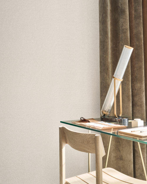 This serene workspace captures the understated elegance of Omexco's Pure collection, reference PU202, titled Resource. The wallpaper showcases a refined weave of twisted paper, artfully blending two different yarns of varying thicknesses and shades. The soft, textured surface subtly reflects the light, adding depth and warmth to the space. A modern glass desk paired with a minimalist wooden chair complements the neutral tones of the wallpaper, while a contemporary lamp with brass accents adds a touch of sophistication. The rich velvet curtains in earthy hues complete the setting, creating a tranquil and inspiring environment.