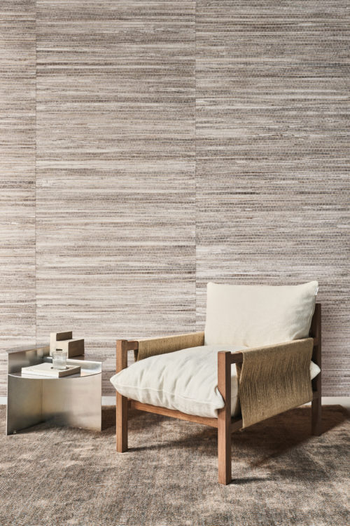 This serene reading corner features Omexco's Pure collection, reference PU222, known as Innate. The wallcovering showcases a refined abaca weave, skillfully crafted in a choice of rich, thick texture or a finer, more delicate variant, both enhanced with a subtle coat of white paint. The natural striations create depth and add a warm, organic feel to the space. A minimalist wooden armchair with cream cushions is paired with a silver metallic side table, holding a stack of books and a glass. The neutral tones of the wall and furnishings complement each other, creating an inviting yet understated aesthetic.