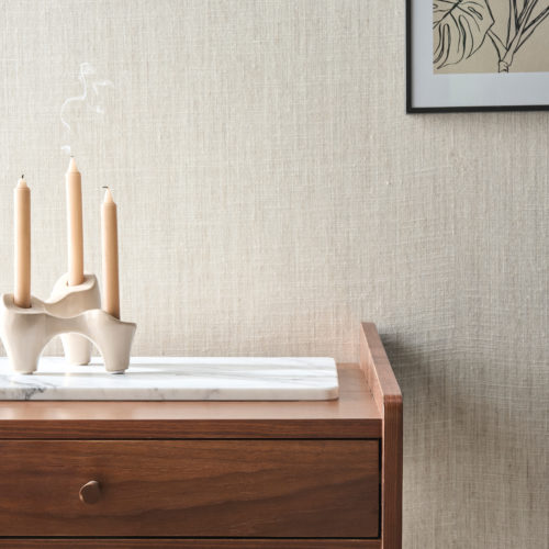 A refined interior showcasing Omexco's Pure collection, reference PU322, a wallpaper that embodies the purity of linen. The natural fibers and textured surface create a serene and timeless aesthetic, perfect for modern and minimalist interiors. The scene features a mid-century inspired wooden sideboard with clean lines, topped with a marble tray holding an artistic white ceramic candleholder with three softly burning beige candles. In the background, a framed botanical artwork complements the natural tones of the wallcovering, enhancing the understated elegance of the space.