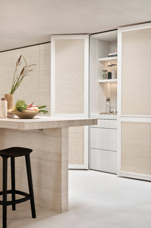 A sophisticated kitchen interior featuring Omexco's Pure collection, reference PU292, a wallcovering showcasing sisal in its most elegant simplicity. The natural texture of the wallpaper adds depth and warmth, creating a serene and refined ambiance. The scene includes a central stone kitchen island with clean, minimalist lines, paired with a sleek black barstool for a touch of contrast. On the countertop, a wooden bowl filled with fresh vegetables and a ceramic vase with dried grasses highlight the harmony between nature and design. In the background, paneled cabinets with integrated shelving and sisal detailing seamlessly blend functionality with aesthetic appeal, offering a perfect balance of practicality and elegance.