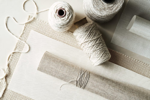 A Pure collection moodboard featuring an elegant composition of natural materials. The image includes woven textures, linen fabric, and multiple spools of thread in soft, neutral tones. A rolled fabric sample wrapped with matching thread is placed centrally, surrounded by delicate yarns and woven samples, emphasizing the tactile richness and craftsmanship of the collection.