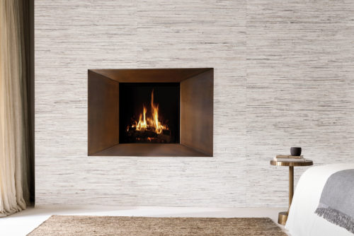 A serene bedroom interior featuring Omexco's Pure collection, reference PU241, known as Native. This elegant wallcovering displays a bold weave of wide abaca strips, expertly coated with white paint, creating a refined yet organic texture. The fireplace, framed in a brushed bronze surround, serves as a striking focal point, exuding warmth and sophistication. Complementing the ambiance, a minimalist bedside table with a brass finish holds a simple cup and book, while neutral curtains and a textured jute rug enhance the natural palette. This design perfectly marries contemporary elegance with raw natural beauty.