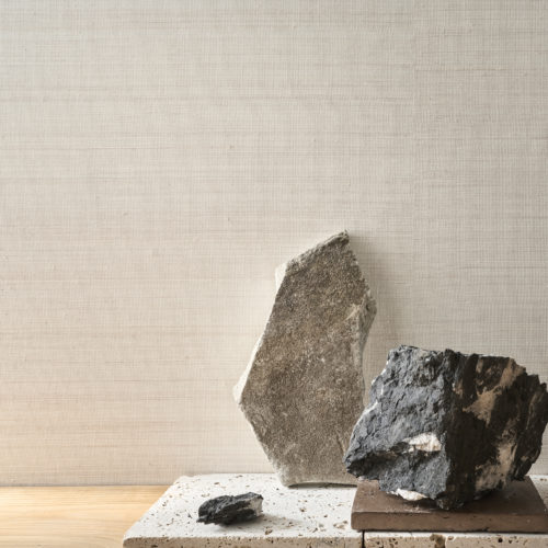 A minimalist composition featuring Omexco's Pure collection, reference PU371, composed of a regular weave of natural-coloured abaca, applied on a white background. The wallcovering’s natural fibers and subtle texture evoke an organic and timeless elegance. The scene highlights an arrangement of raw stone pieces in various shades of grey and beige, placed on textured and polished bases, creating a balance between raw natural elements and refined design. The neutral tones of the wallcovering provide a serene backdrop, complementing the earthy materials and minimalist aesthetic.