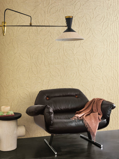 This elegant reading nook features the OAS222 reference, titled Verano, from Omexco’s Oase collection. The wallcovering is crafted from exquisite paperweave, embroidered with a subtle organic pattern that adds depth and refinement. The warm, neutral tones of the wall harmonize beautifully with the luxurious black leather armchair, which is adorned with a soft, fringed throw for added comfort. A sculptural side table with a raw, industrial aesthetic enhances the space’s unique character. Overhead, a vintage-inspired wall lamp with brass accents casts a soft glow, completing the scene with a touch of mid-century sophistication.