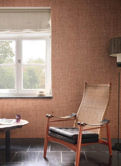 This inviting interior features the OAS294 reference, titled Cesta, from Omexco’s Oase collection. The wallpaper is crafted from paper fibres dyed with a gentle irregularity, evoking the natural textures and subtle variations found in organic materials. The warm, earthy hues create a serene and grounding atmosphere, harmonizing beautifully with the surrounding decor. A woven rattan chair with a rich wooden frame enhances the natural aesthetic, while a sculptural side table made of raw wood adds a touch of artisanal craftsmanship. The neutral-toned linen curtains softly diffuse the daylight, reinforcing the calm and inviting ambiance of this space.