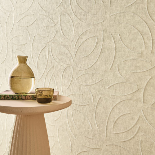 This serene interior highlights Omexco’s OAS221 reference, Verano, from the Oase collection. The wallcovering, crafted from exquisite paperweave, features delicate embroidered patterns inspired by organic forms, adding a sense of movement and sophistication to the space. The soft, neutral tones create an inviting and calming atmosphere, perfectly complemented by a sculptural side table, a glass water carafe, and a textured ceramic cup. The interplay of light and shadow on the embossed surface enhances its depth, making it a refined choice for elegant and natural interiors.