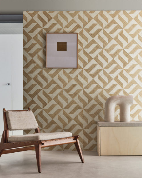 A captivating decor shot featuring an ethnic Scandinavian-style wooden chair on the left and a light wood cupboard on the right adorned with a ceramic decorative element. The grey cast floor provides a neutral foundation, while the backdrop showcases Omexco's Intuition wallcoverings—a raffia patchwork displaying a striking geometric design with yellow and white shapes, creating a harmonious and stylish interior composition.