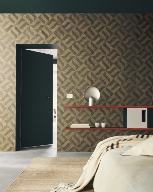 A serene bedroom with a grey cast floor, featuring a green open door on the left. Wall shelves adorn the room, hosting a grey metal minimalist table lamp. The bed is dressed in beige sheets, complemented by a matching beige carpet. The backdrop showcases Omexco's Intuition wallcoverings—a raffia patchwork with an appealing geometric design in green and light brown shapes, creating a calming and stylish atmosphere.