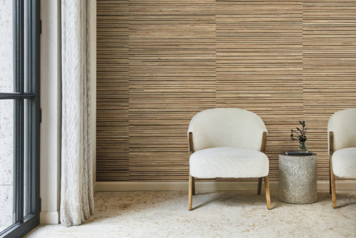 Interior decor shot featuring a stone tile floor adorned with two bouclé fabric white armchairs, each with a wooden frame. A cylinder-shaped side table is positioned between the chairs. A large window with linen curtains is on the left. The backdrop showcases Omexco's handmade Intuition wallcoverings in shades of light and dark brown, crafted with meticulous reliefs using natural fibers such as banana bark, banana stem fiber, and water lily.