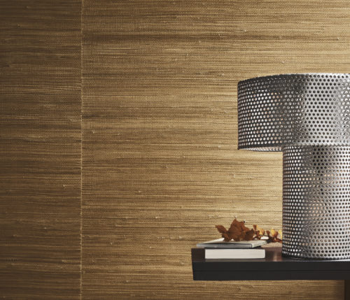 This closeup image showcases a black side table positioned against a wall. Resting atop the table is a sleek metal table lamp with small perforations in it. Next to to the lamp, two books are neatly stacked, accompanied by a twig adorned with autumn leaves, adding a touch of seasonal charm to the scene. In the background, Omexco’s natural handmade Intuition wallcoverings serve as a backdrop. This wallcovering features a minimalist weave, in orange-brown, highlighting the inherent beauty of youthful abaca skin fibre.