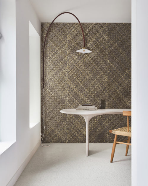This study space features a white floor and a white wall on the left, illuminated by natural daylight streaming in through two windows. In the center of the room sits a sleek organic-shaped desk paired with a rattan chair, creating a harmonious blend of modern and natural elements. On the desk, there are several books neatly arranged. The focal point of the room is the impressive wall showcasing Omexco's handmade natural wallcoverings collection "Intuition". The wall is adorned with textured woven pandan (basket weave) in shades of brown, adding warmth and texture to the space.