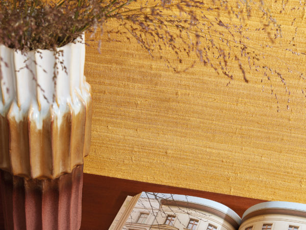 Closeup of an open architecture book laying next to a tricolor ceramic 70's vase. The vase is holding some dried flowers. Both items are placed on a wooden table or sideboard, standing against an ochre wall with a precious silk textile design. These are Omexco’s "Loom Stories” wallcoverings, a rich and eclectic collection of irregular textures made from hand-spun yarns, fine Belgian linen and wild silk, all in a diverse palette that ranges from soothing neutrals to eye-catching, vivaciously bold colours.