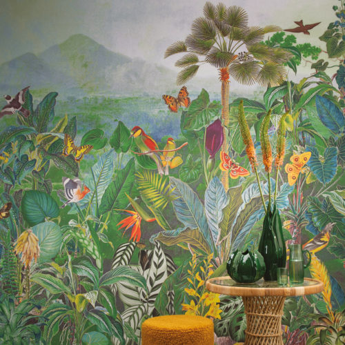 Closeup of a large mural jungle print showing exotic plants, paradise birds, palm trees and butterflies. This four drop panoramic deisgn from Omexco’s "Birds of Paradise” collection is printed on non-woven wallcoverings and immerses us in nature’s splendour. In front of the wall, we see a round ochre pouf as well as a ratan side table on which two green ceramic vases are posed.