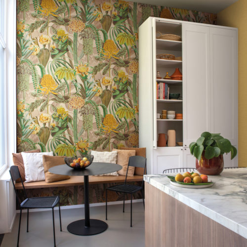 Example of a modern kitchen with flowery botanical wallcoverings and a cosy breakfast nook next to the window. Shown here are Omexco’s exuberant “Birds of Paradise” wallcoverings, a fabulous foliage in shades of yellow, lime green, khaki and beige. The inviting kitchen also has a kitchen island with a marble tablet and a large white pantry.