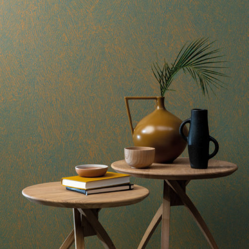 Contemporary mini decor with two wooden side tables and ceramic, organic shaped vases in ochre and black posed on top of them. The wall is decorated with printed non-woven wallcoverings by Omexco. The design, shown here in shades of teal and ochre, is inspired by a special painting technique that creates a brushed chalk effect on the wall. The “Chalk” design is one of the five designs of the High Performance Patina Wallcovering collection.