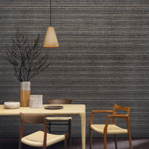 The Atelier collection by Omexco is an eclectic blend of artisanal craftsmanship and contemporary couture. On the wall of this inviting dining room we see a horizontally shown chunky weave in dark grey, brown and beige hues. The space is furnished with a Scandinavian style dining table and chairs, marked by a focus on clean, simple lines and minimalism. We also see a a chestnut parquet floor, a hanging lamp with wooden lightshade and ceramic vases and plates on the table.