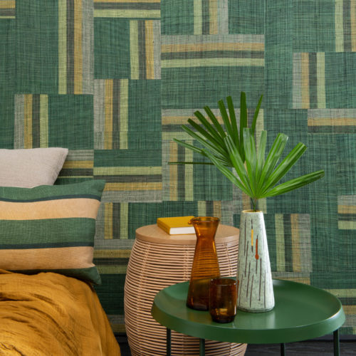 Authentic craftmanship embodies the bold Atelier collection designed by the creative minds of Omexco. This image shows an impressive green patchwork of handwoven raffia. The perfect statement wall in this tropical bedroom with ochre colour accents, a green metal bedside table, exotic plant leaf and cosy bed pillows.