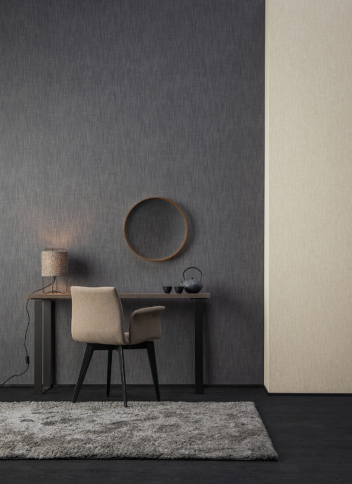 Hotel room where Omexco's High Performance Textures wallcoverings are installed. We see a desk with chair, round mirror, fluffy carpet and contrasting walls in dark grey and beige. Denim design, luxurious yet sustainable. Inspired by the technique used for jeans manufacturing with diagonal ribbing as a result.