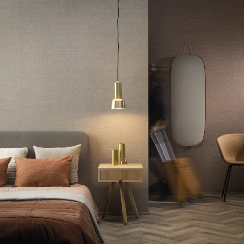 On the walls of this hotel room: the Abaca design in beige and blush hues from Omexco's High Performance Textures collection. It's a semi plain non-woven wallcovering with an intriguing texture, shown horizontally. The wallcovering is paired with a cosy bed, pendulum lamp, orange and beige pillows, orange chair and a wooden side table.