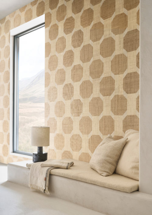 A serene corner featuring a built-in bench adorned with soft beige cushions and a fringed throw, set against a striking wallcovering from Omexco's Pure collection, reference PU431. The wallcovering showcases a vibrant and sophisticated marquetry of raffia, with a bold geometric pattern in natural tones that adds depth and texture to the space. To the left, a sleek, minimalist black table lamp with a cylindrical beige shade sits atop a modern concrete ledge, its clean lines complementing the natural materials. The large window frames a picturesque view of rolling hills, bringing a touch of the outdoors into this elegant and harmonious interior.